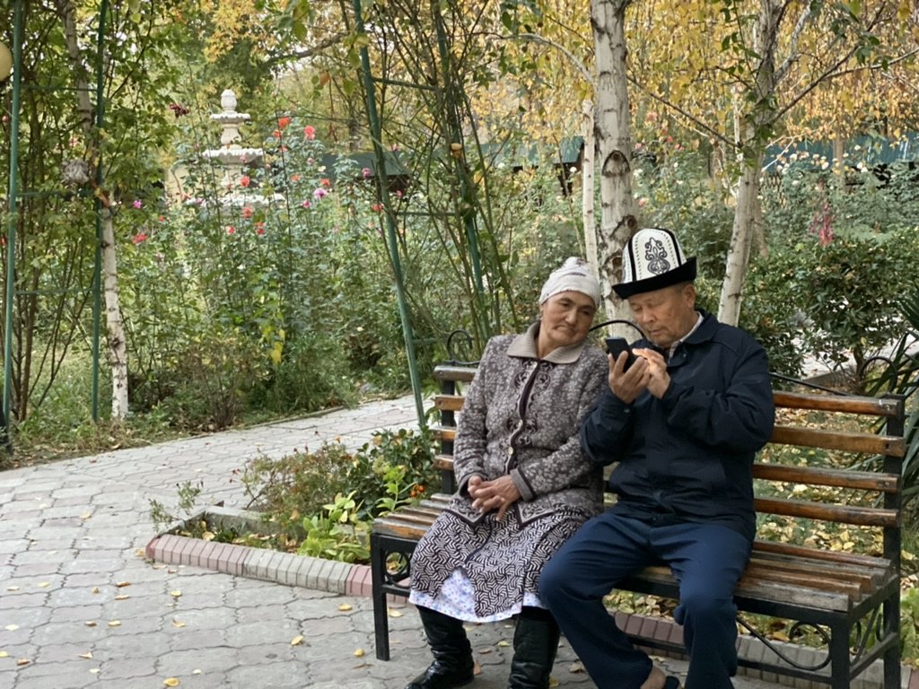 An elderly Kyrgyz couple sitting on a park bench looking at a mobile phone