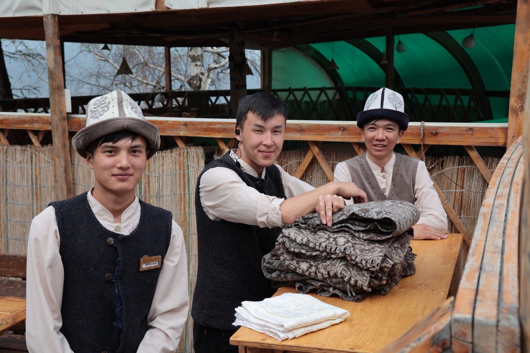 Three young Kyrgyz men in traditional kalpak hats with handmade felt shyrdak rugs
