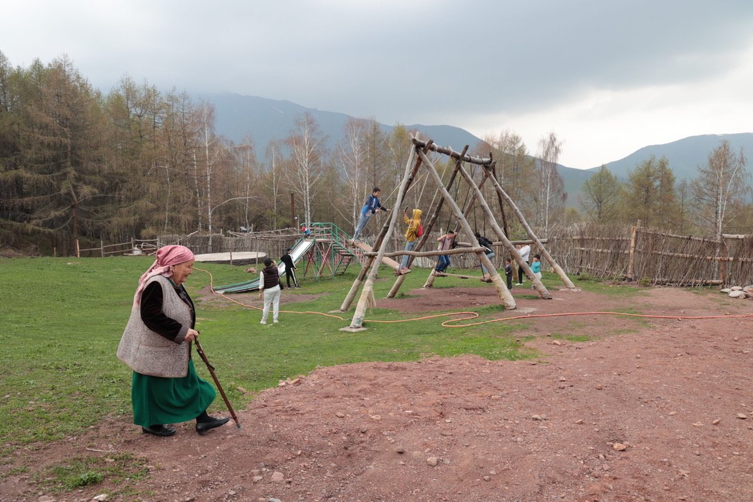 An elderly woman walking with a stick past children playing on swings in a mountain village